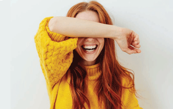 Image of a smiling teenage girl in a yellow jumper with her arm raised over her eyes