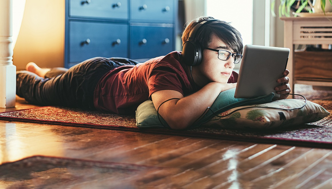 Teenager lying on the floor with headphones and watching something on a tablet.