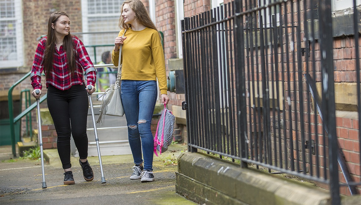 A young woman helps her friend who is on crutches by providing support and carrying her bag.