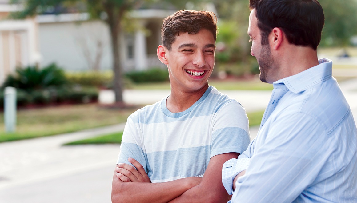 A father and his 13 year old teenage son standing in their front yard, smiling at each other with arms crossed.